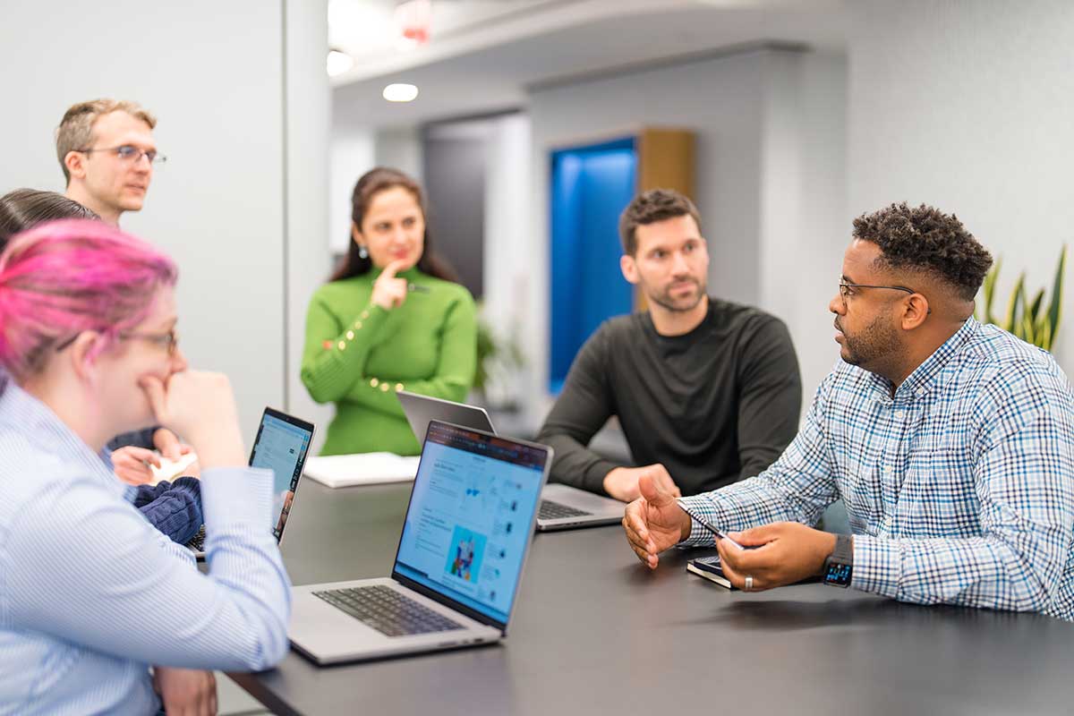 Six people have a discussion during a course at Khoury College in Arlington. Four students are seated around a large table and two other people are standing at the far end