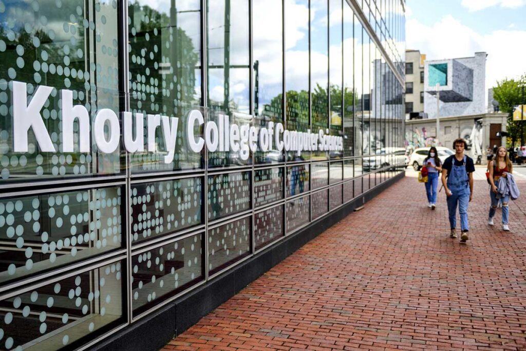 Three students walk on the brick sidewalk alongside West Village H, which has a Khoury College of Computer Sciences sign on the exterior