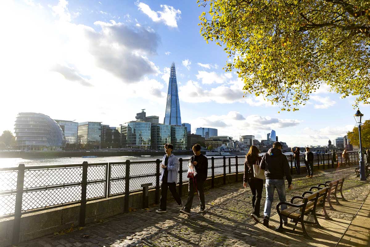 People walk along the Thames River in London as the sun sets behind the river. In the background, a large skyscraper is visible.