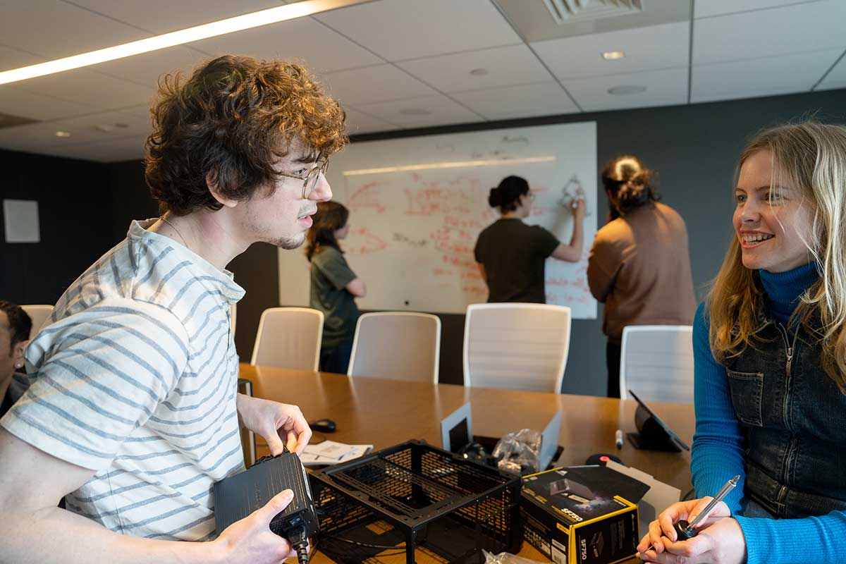 Two students discuss their research while standing on opposite sides of a table. A black device is on the table betwen them, and three students are writing on a whiteboard in the background.