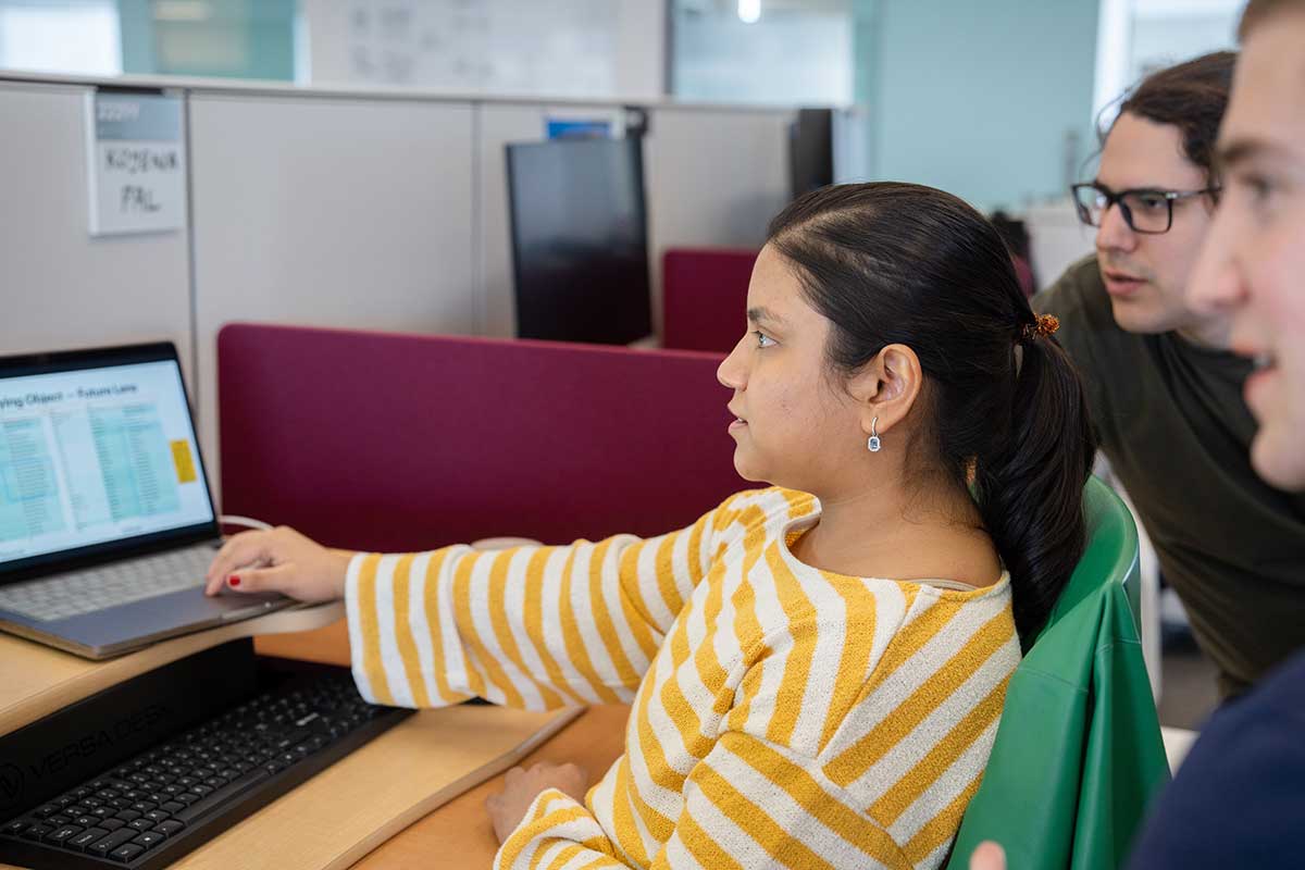 A student types on a laptop while sitting in a chair. Two colleagues view the screen while leaning forward.