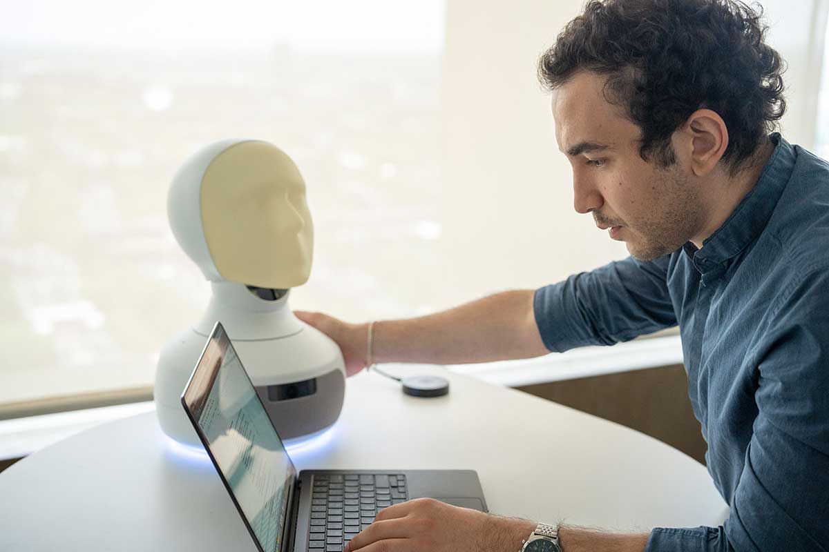 A student types on a laptop with one hand while grasping a device shaped like a human head and neck.