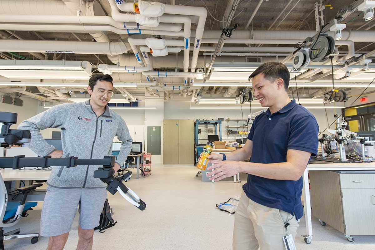 A student stands in a robotics lab while holding a bottle of juice in front of a robotic arm that appears to be reaching for the bottle. A second student watches the robotic arm.