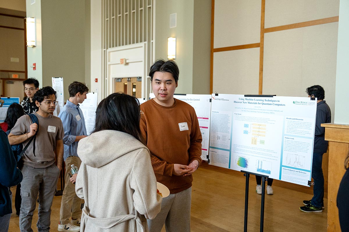 A student explains a research project to another student during a research expo. The student is standing to the left of his research poster.