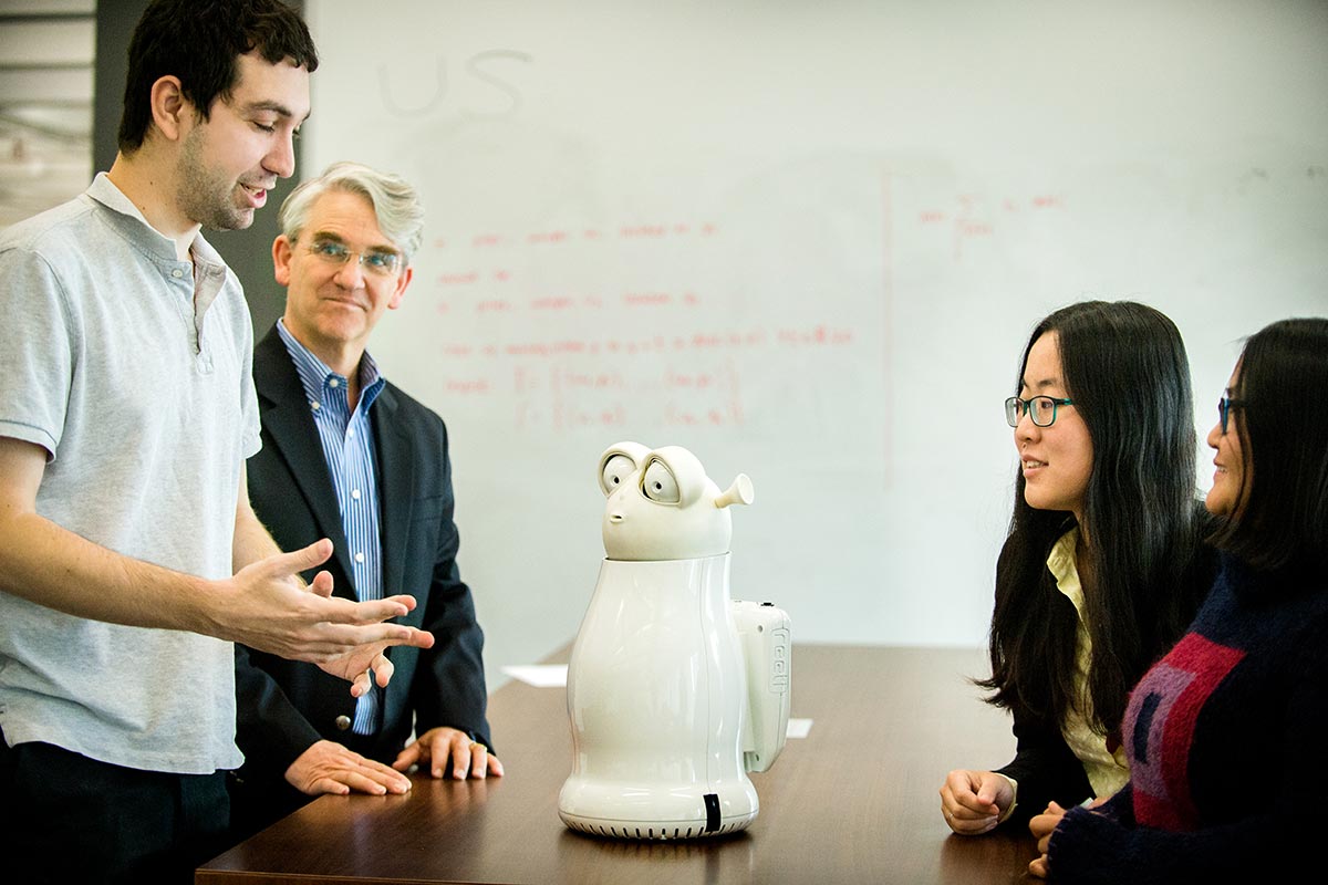 Four people stand around a table on which a personal health robot stands. The robot is white, with two large eyes and a small mouth. Two people stand on the left side of the table, and the other two stand on the right.