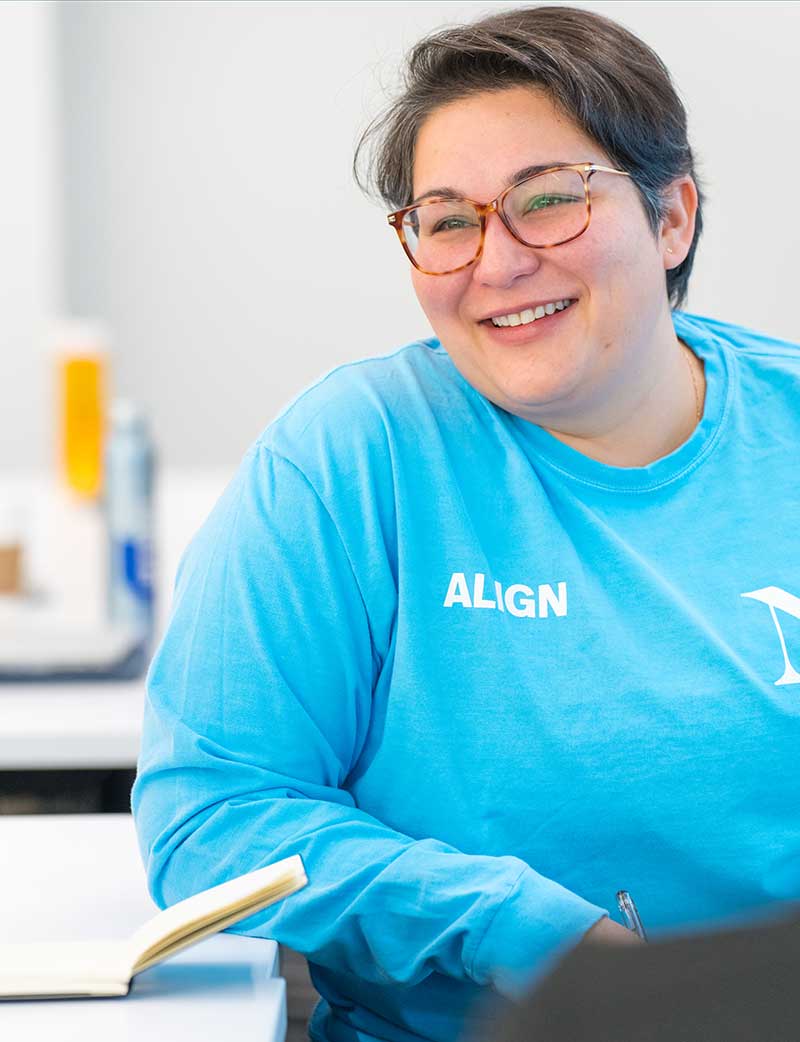 A student sitting at a large table smiles during a presentation. An open notebook sits on the table in front of the student.