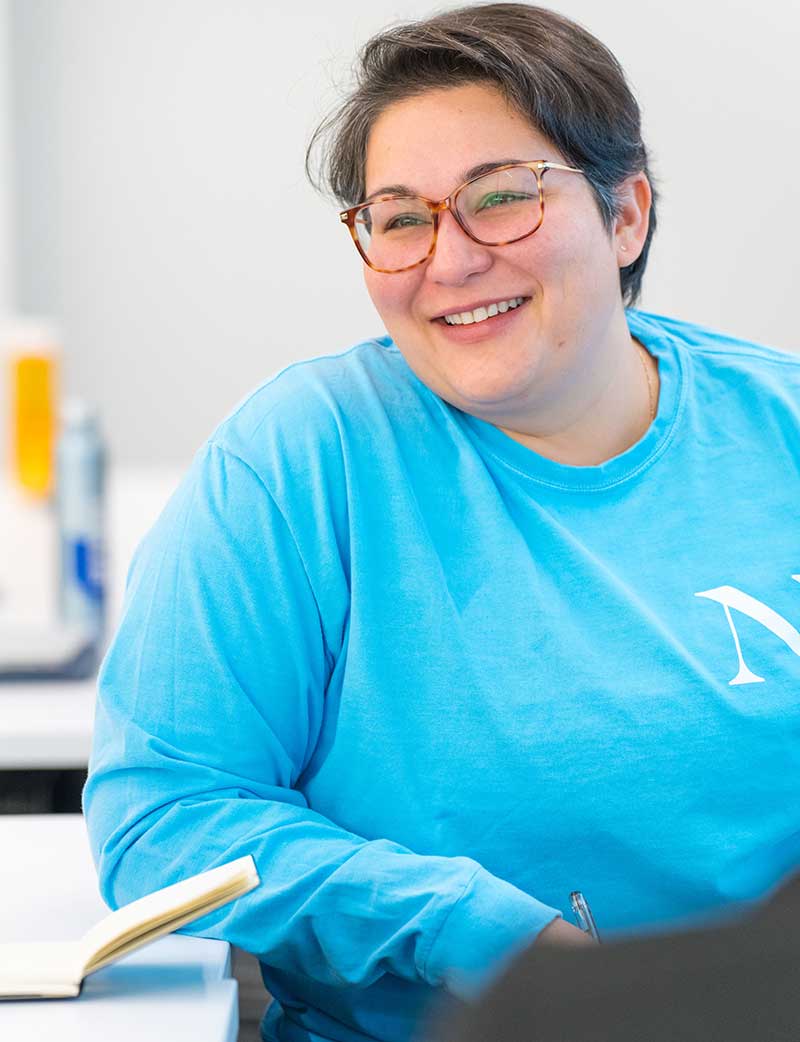 A student sitting at a large table smiles during a presentation. An open notebook sits on the table in front of the student.