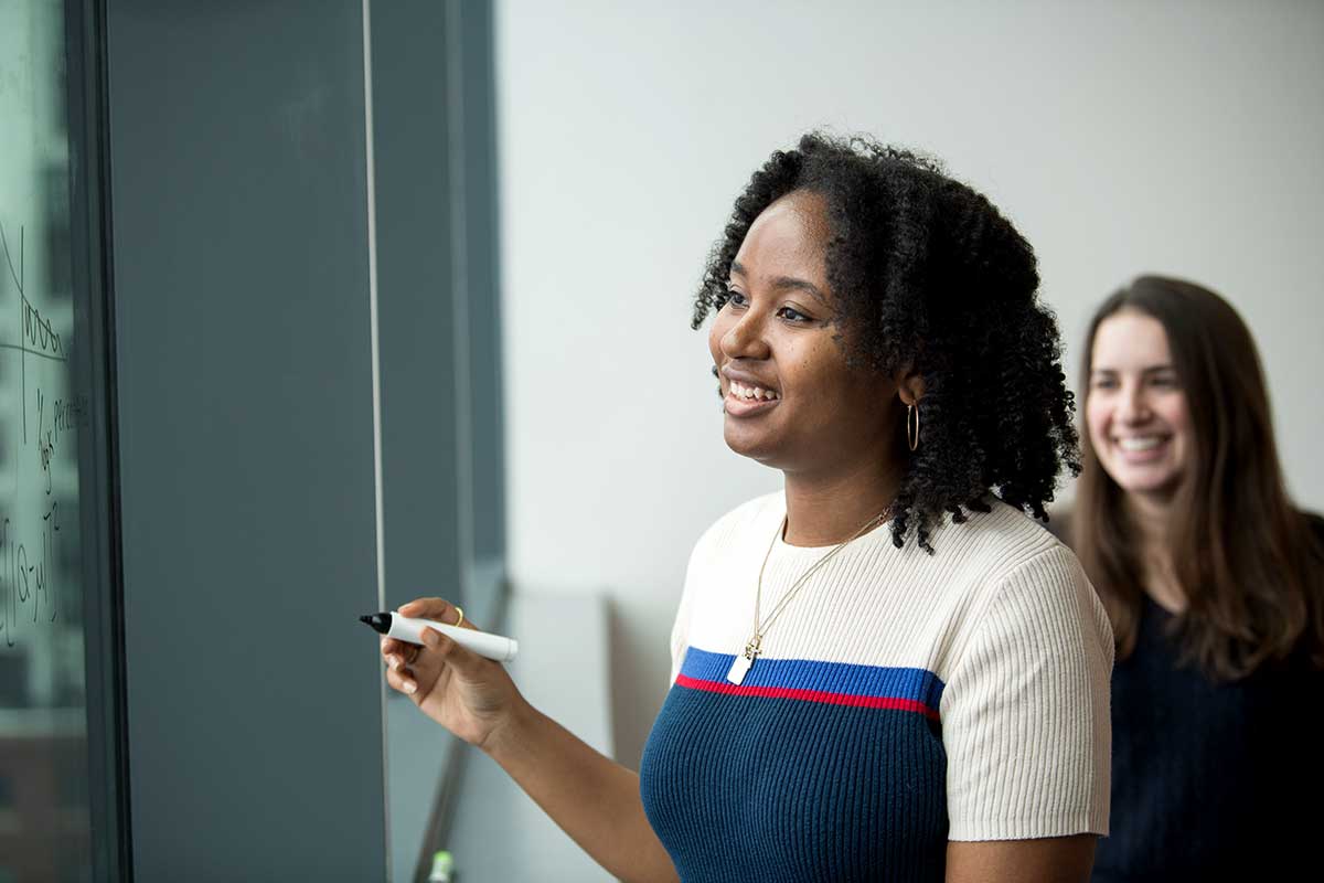 A Khoury student holds a marker while looking at symbols drawn on a smart board in a Khoury classroom. In the background, another student smiles.