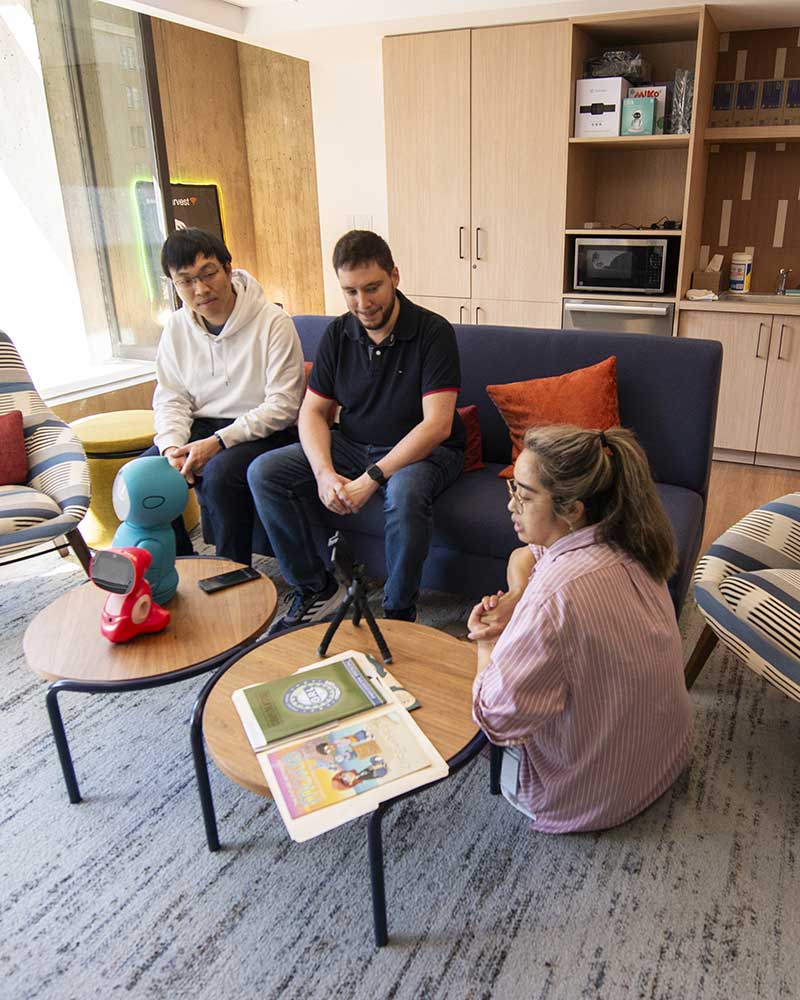 Three cybersecurity researchers sit around a set of tables with various wireless devices. Two students are sitting on a couch and the other is sitting on the floor.