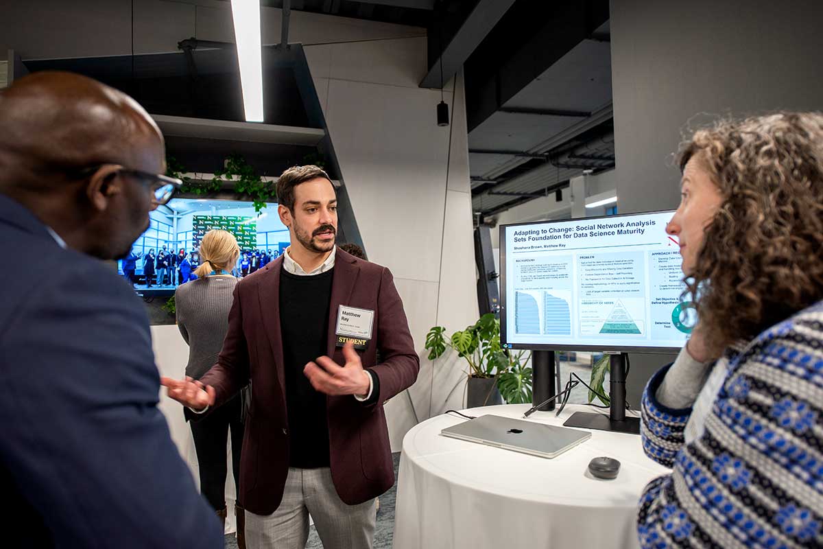 A student explains a research project to two faculty members during a research showcase. The student's research poster appears on a computer display propped on a circular table.