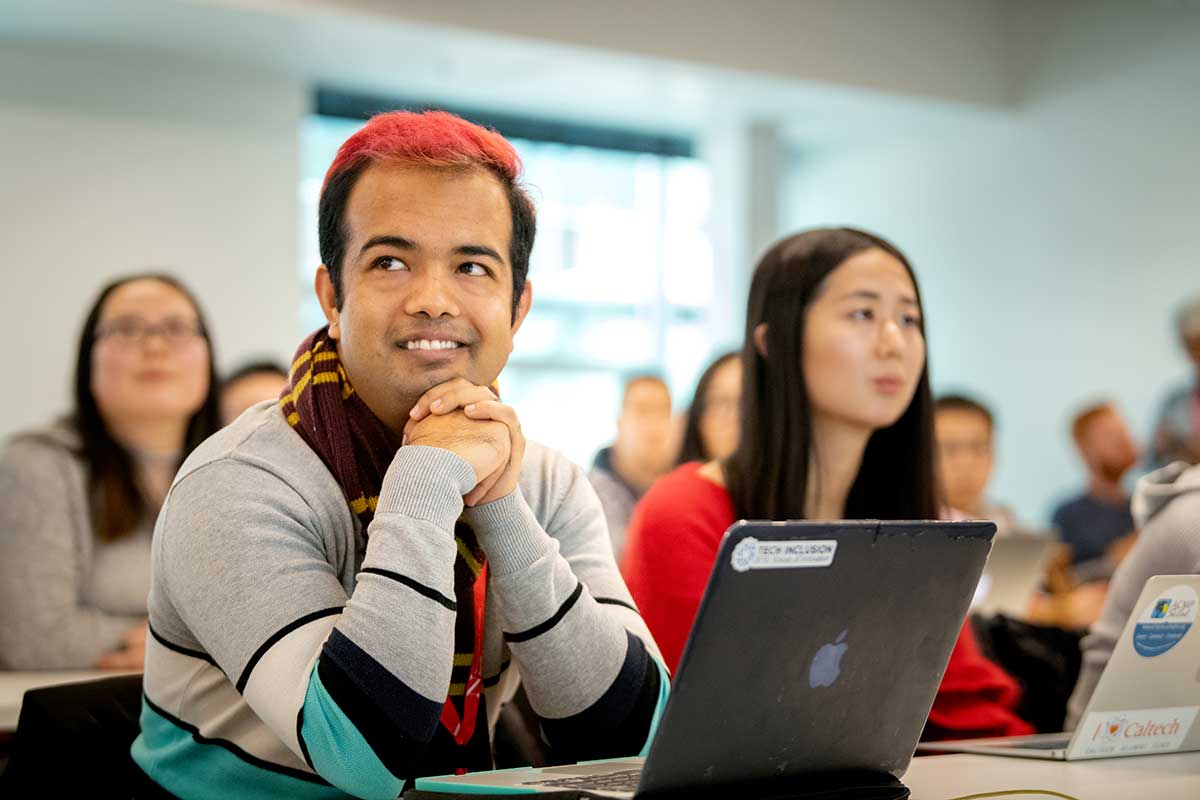 Several rows of students site at tables and view a course lecture. The student closest to the camera smiles with a laptop open on the desk.