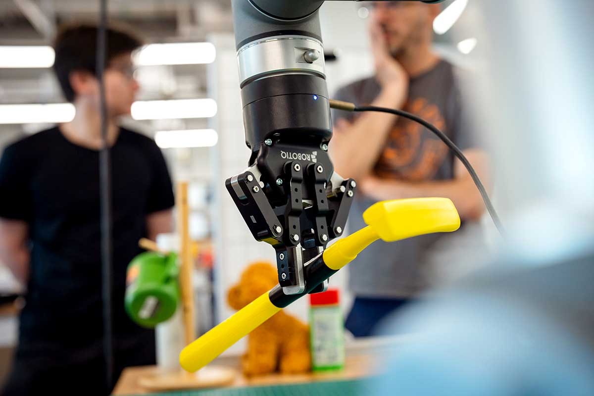 A robotic arm with a grasping mechanism at the end of the arm grasps a yellow plastic object shaped like a hammer. Two students look on in the background.