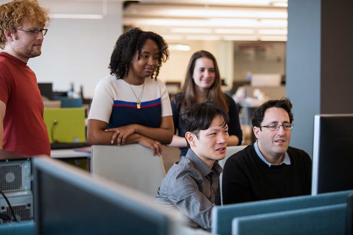 Khoury faculty member Daniel Wichs, right, looks at a computer screen while sitting at a desk next to a student, who is also looking at the screen. Three students stand behind them.