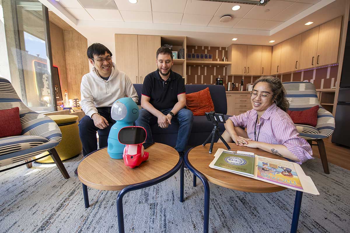 Three students laugh during a conversation in a Khoury research lab. Two of the students are sitting on a couch while the student art right sits on the floor while she speaks into a device attached to a tripod.