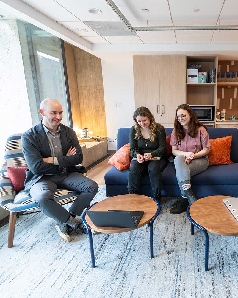 Khoury faculty member Christo Wilson, left, sits in chair while two students sitting on a couch view a laptop screen.