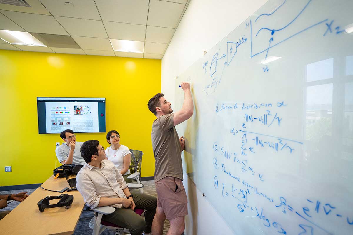 A student uses a blue marker to write on a whiteboard in a classroom while other students look on