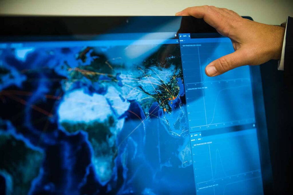 A hand rests on a large computer monitor that is showing an info graphic that contains a world map with several bright red lines leading to red dots.