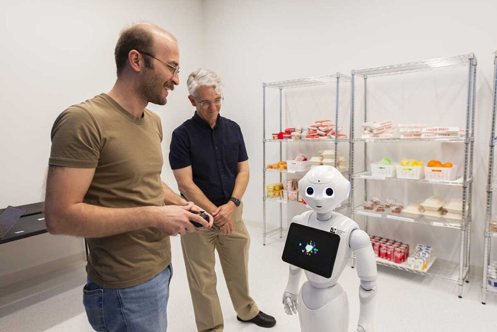 Khoury faculty member Tim Bickmore, right, smiles as a Khoury researcher holding a video game controller demonstrates how to control a white, humanoid robot