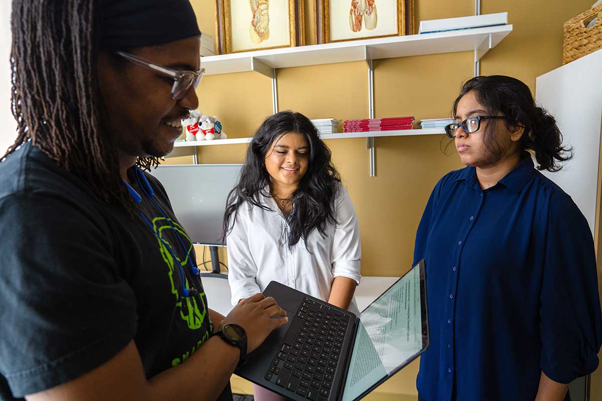 Khoury faculty member Aarti Sathyanarayana, center, discuss research with two students. The student at left is holding an open laptop that shows a research article.