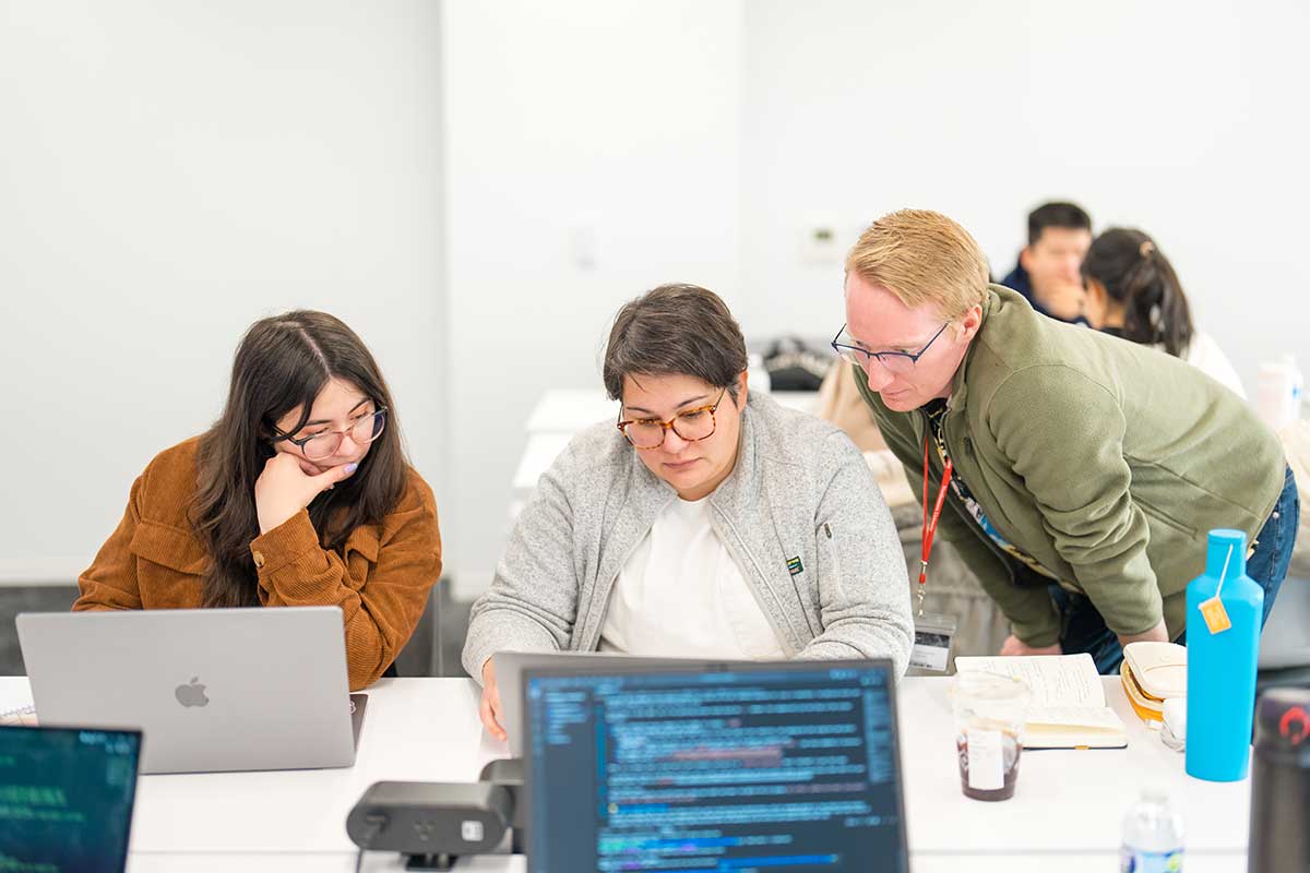 Two students sit at a table and view a laptop screen during a seminar at Khoury College. A faculty member to the student's right is standing looking at the laptop.