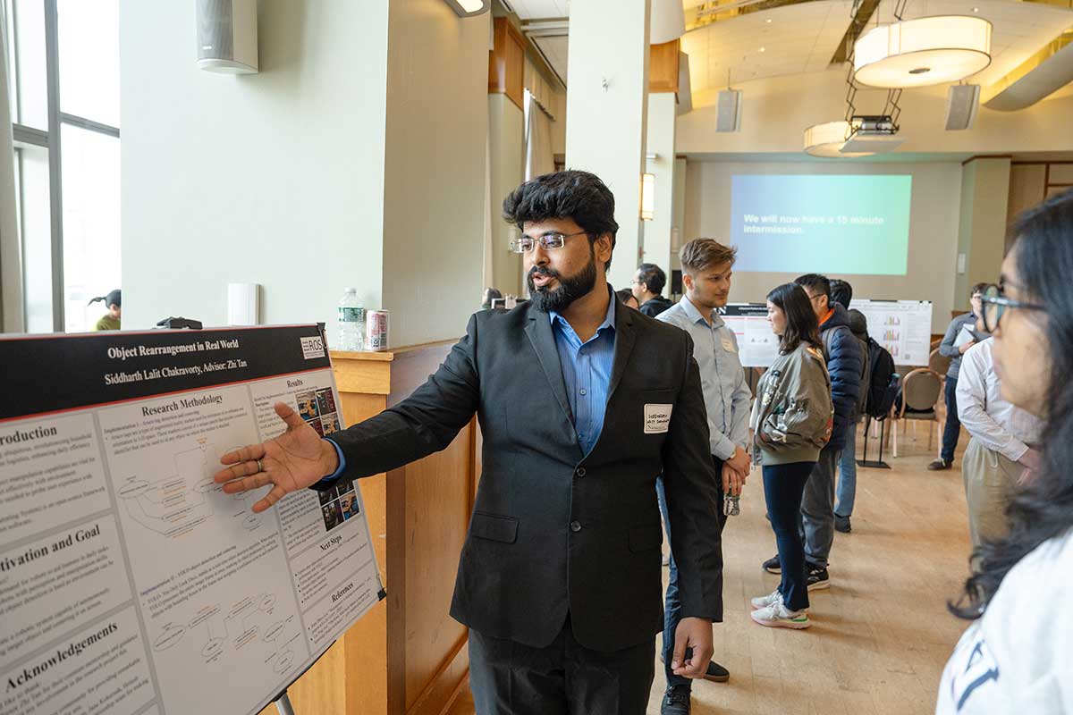 A student standing to the right of his research poster points at the center of his poster as he explains his project to an onlooker. Other students and research posters are in the background.