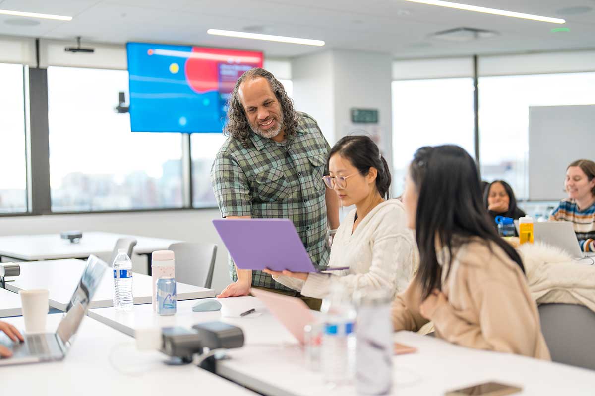 Two students sitting at a table in a lab talk to a Khoury faculty member. The student at left is holding a purple laptop in her hands. The faculty member is standing next to the table looking at the laptop.