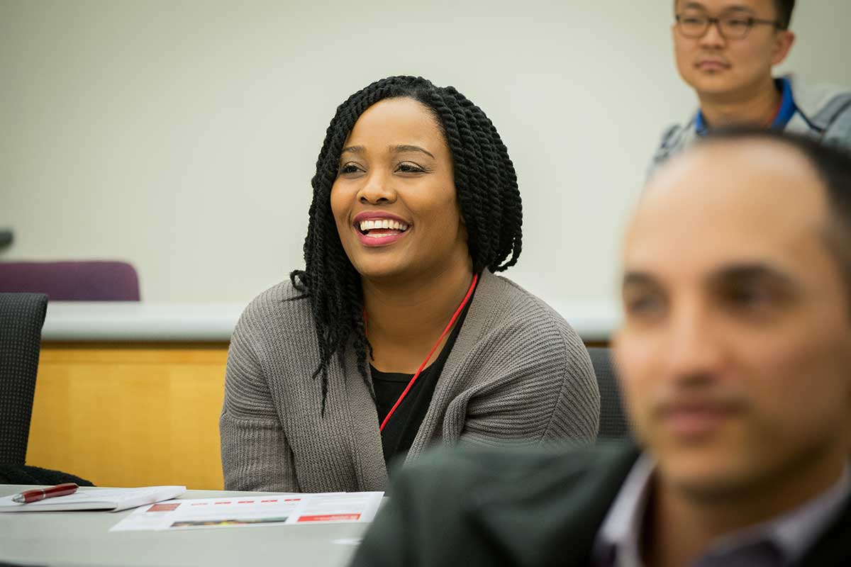 A student sitting in a classroom laughs during a class presentation. Other students are visible in the foreground and background.