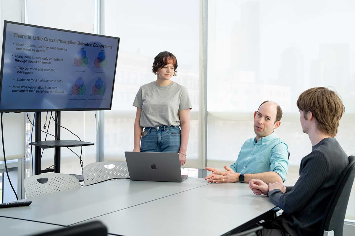 Khoury faculty member Jonathan Bell (center) sits in a chair at a large table while talking to a student (right). Another students stands next to Jonathan. The group is discussing a presentation slide projected on a large screen.