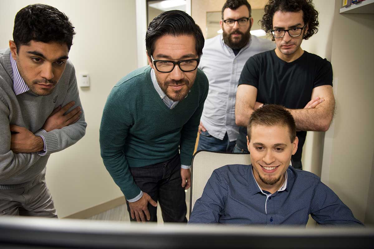 Khoury faculty member Wil Roberston (second from left) views a computer screen along with four other Khoury researchers in a small lab. One students is sitting in a chair in front of the desk; the other four people are standing.