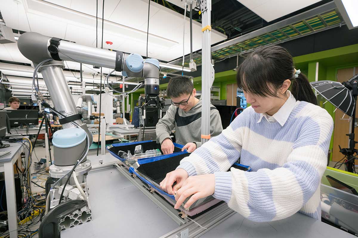 Two students work in the Khoury Robotics Lab. The student on the right is standing behind a table assembling tools and the student on the left is placing objects inside a container.