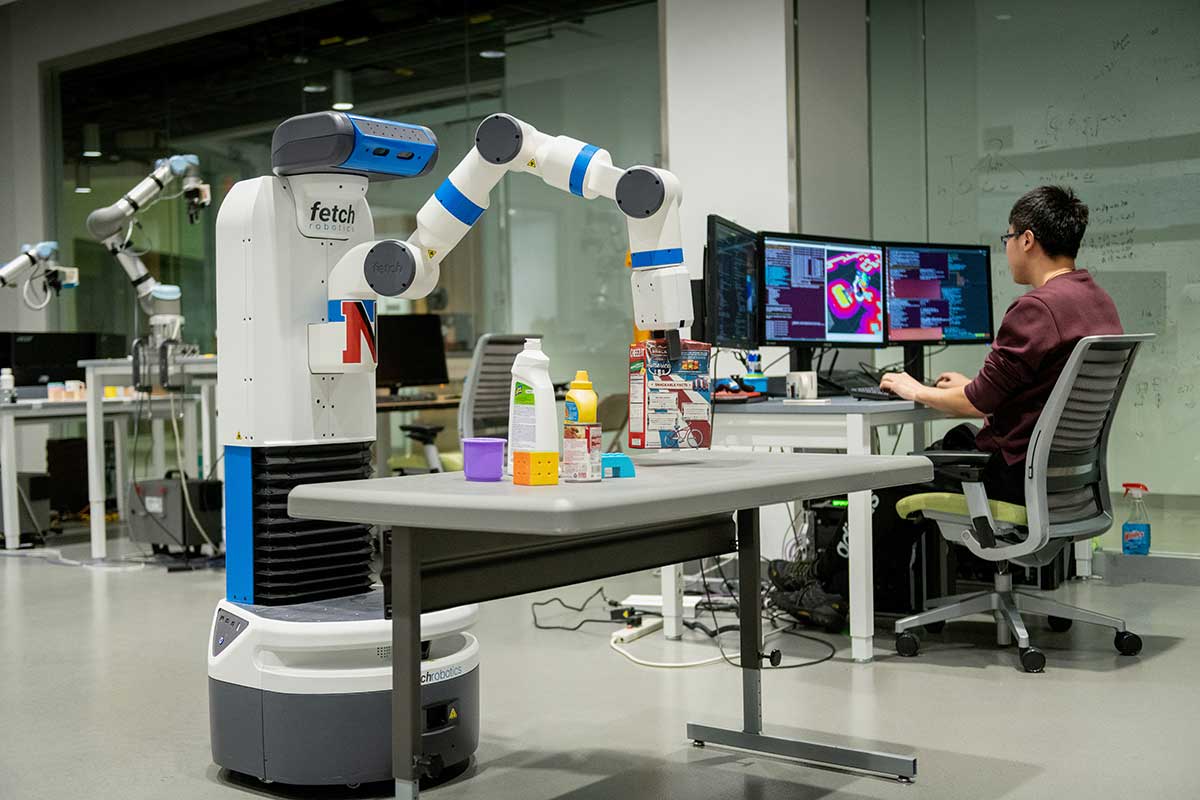 A student sits at a desk typing on a keyboard while viewing three computer monitors. The student is controlling a robotic arm that is lifting a box of cereal off a table.