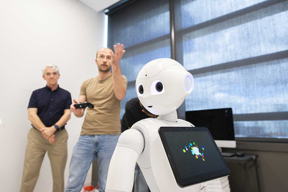 Khoury faculty member Tim Bickmore, left, stands in the background watching another research, who is holding a game controller, to control a white robotic figure.