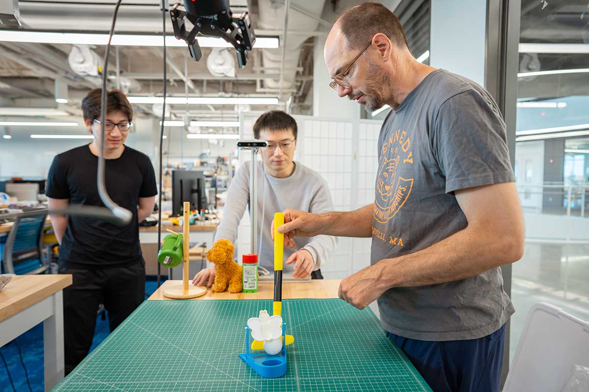 At a drafting table, Khoury faculty member Robert Platt, right, uses a toy golf club to push a ball toward a toy hole. Two students stand on the other side of the table.