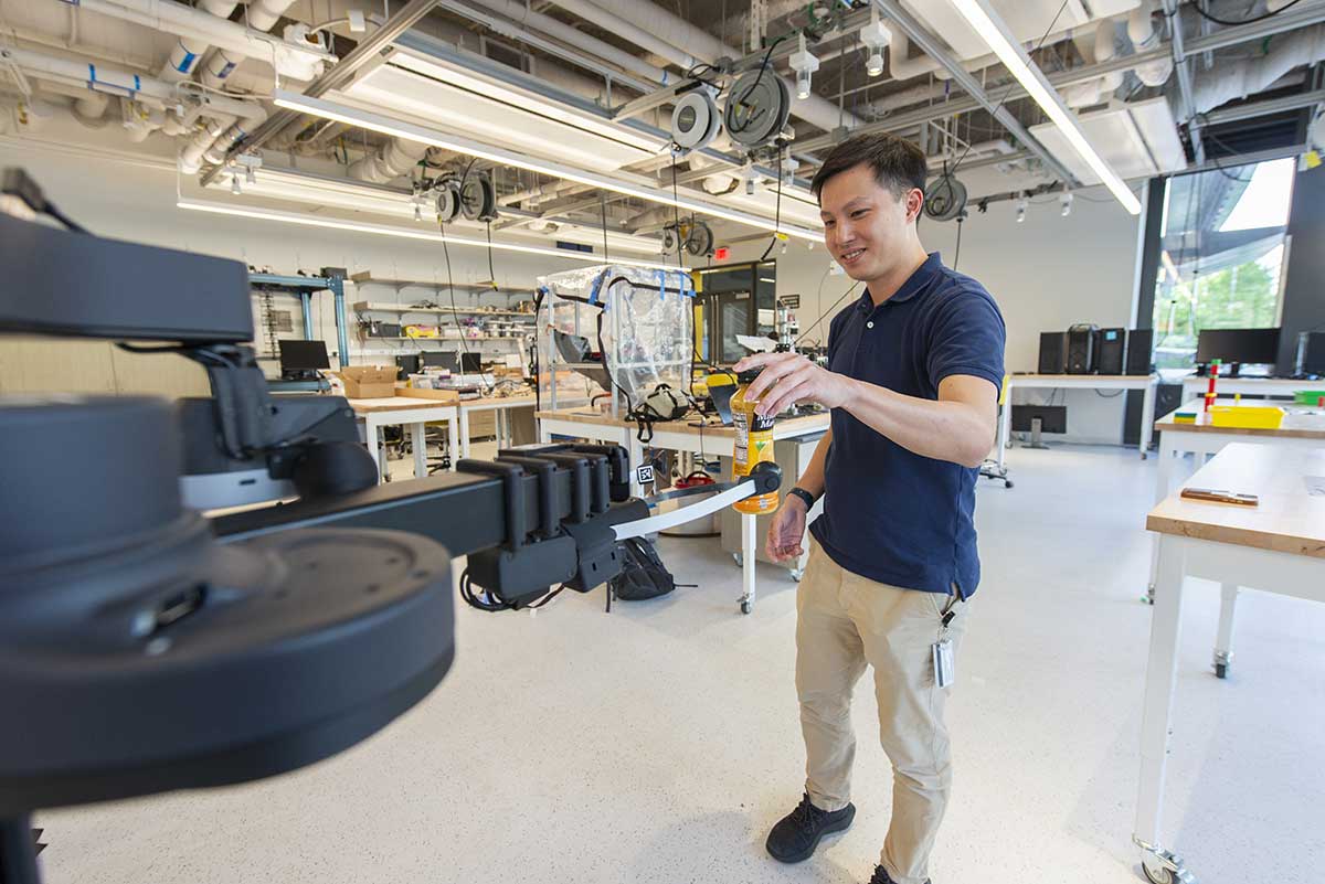 A Khoury researcher standing in a lab attempts to hand a bottle of orange juice to a robotic arm