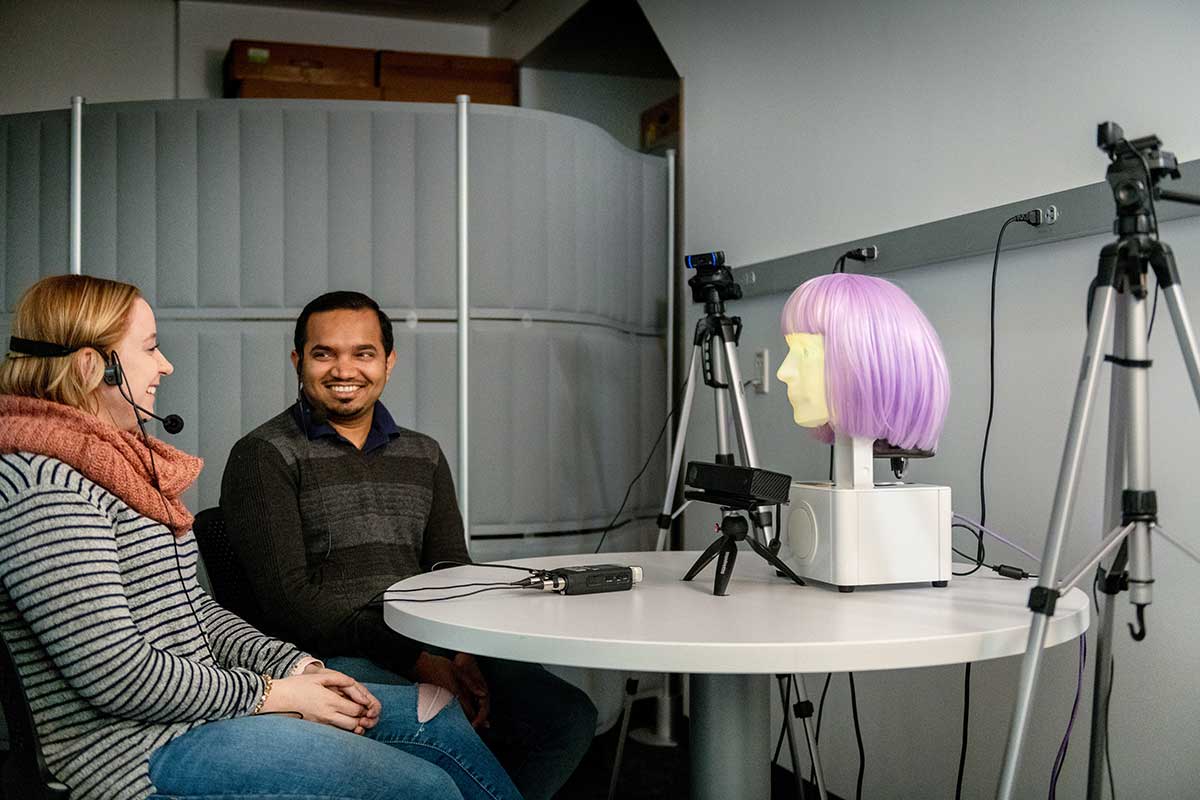 Two students sitting at a table wearing headsets converse with a device that resembles a human face with a purple wig