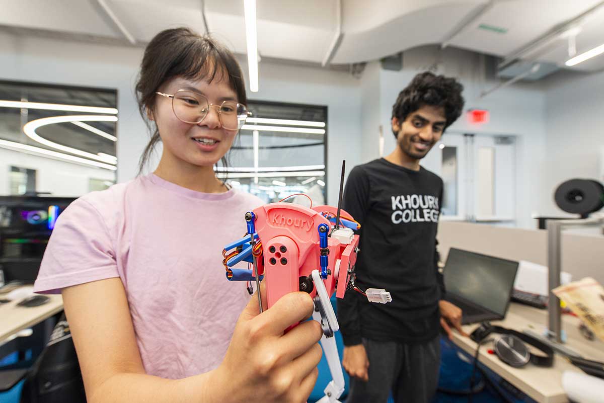 Two Khoury students stand in the Robotics lab. The student on the left is holding a small robotic device with a wire sticking out.