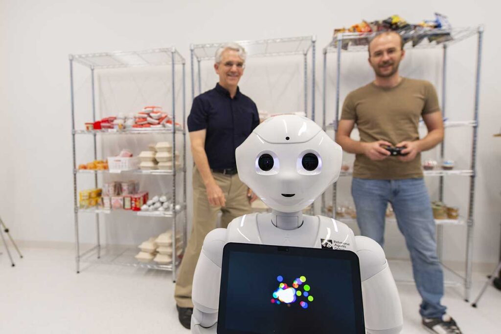 In the foreground, a white robot designed to resemble a human faces the camera with two researchers viewing the robot in the background.