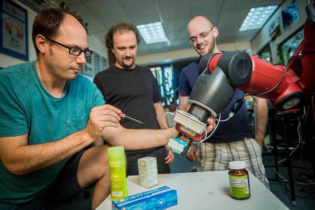 Khoury faculty member Robert Platt, at left, holds a robotic device in his left hand and a screwdriver in his right hand. Two other researchers watch him work.