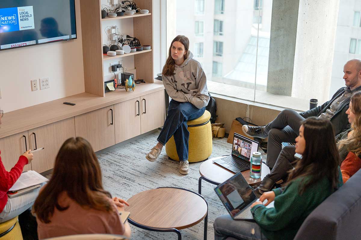 Professor Christo Wilson (right) sits in a lab with a group of students seated on a sofa and stools in a Khoury research lab. In the background, there are several smart devices sitting on bookshelves.