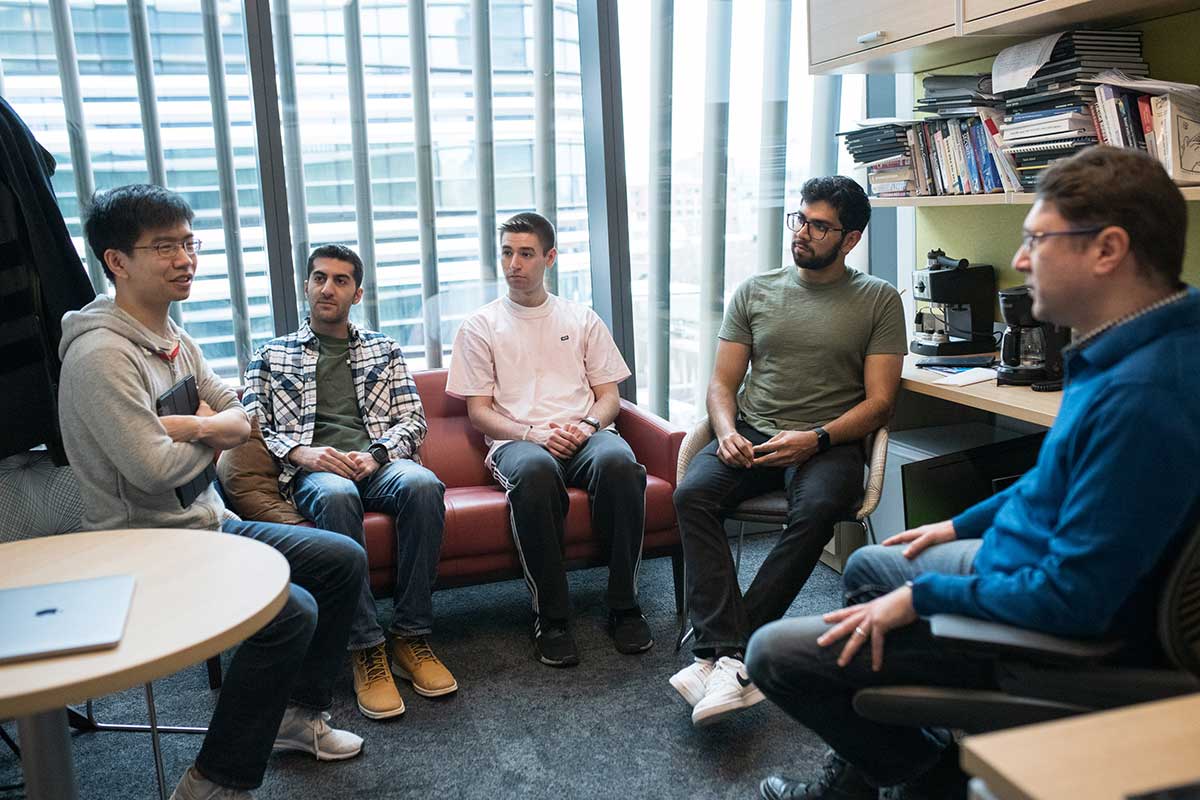 Four Khoury researchers and one Khoury faculty member (right) sit in the faculty member's office as they discuss his research. Three people are sitting in chairs and the two others are sitting on a red sofa.