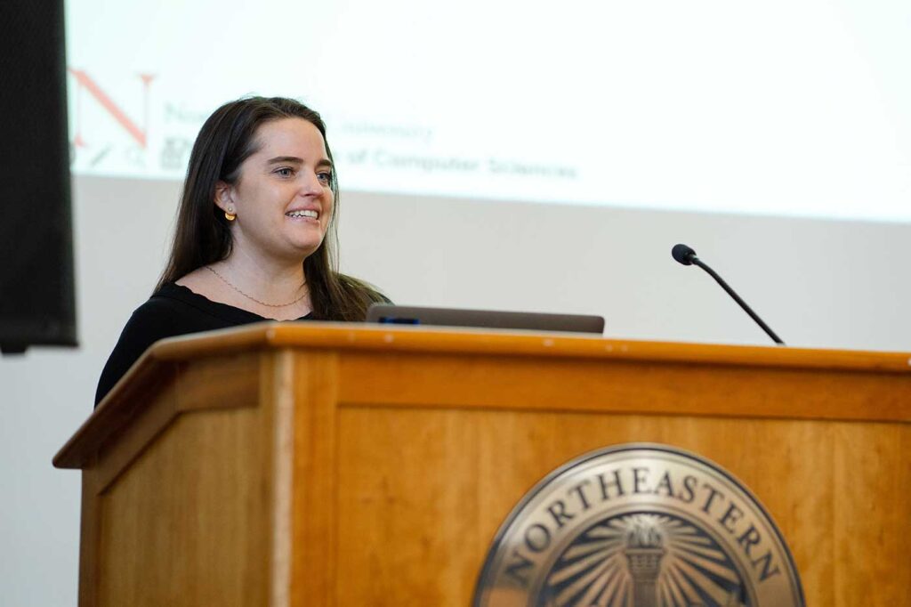 Annie Pates presents a project while standing behind a podium, with the projection screen visible in the background