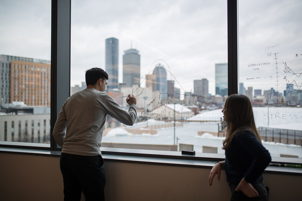 A student writes on a clear window with the Boston skyline visible outside