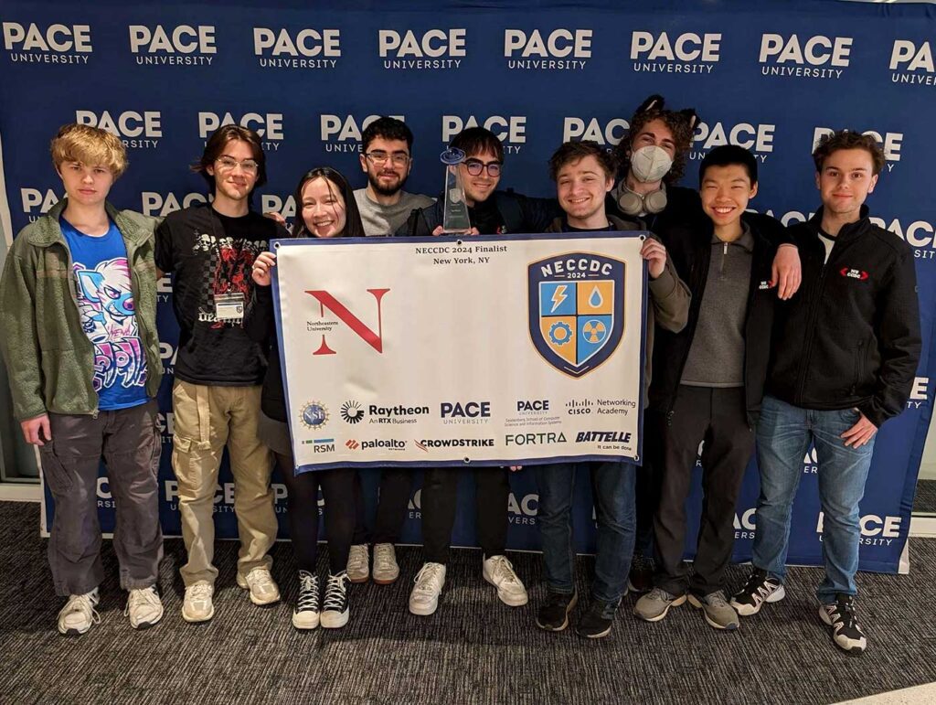 The nine members of Northeastern University’s Collegiate Cyber Defense Competition team pose with a banner and trophy. Two students are holding a NECCDC 2024 Finalist banner, and one student is holding a glass trophy.