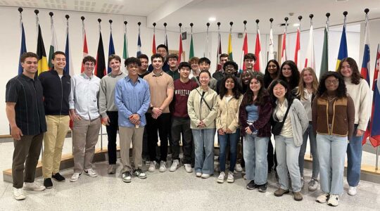 25 students who took part in a Dialogue of Civilizations program in Belgium pose for a photo standing in front of a row of several flags.