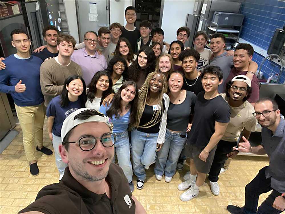 A student takes a selfie of a group of over 20 students and faculty standing in a large kitchen.