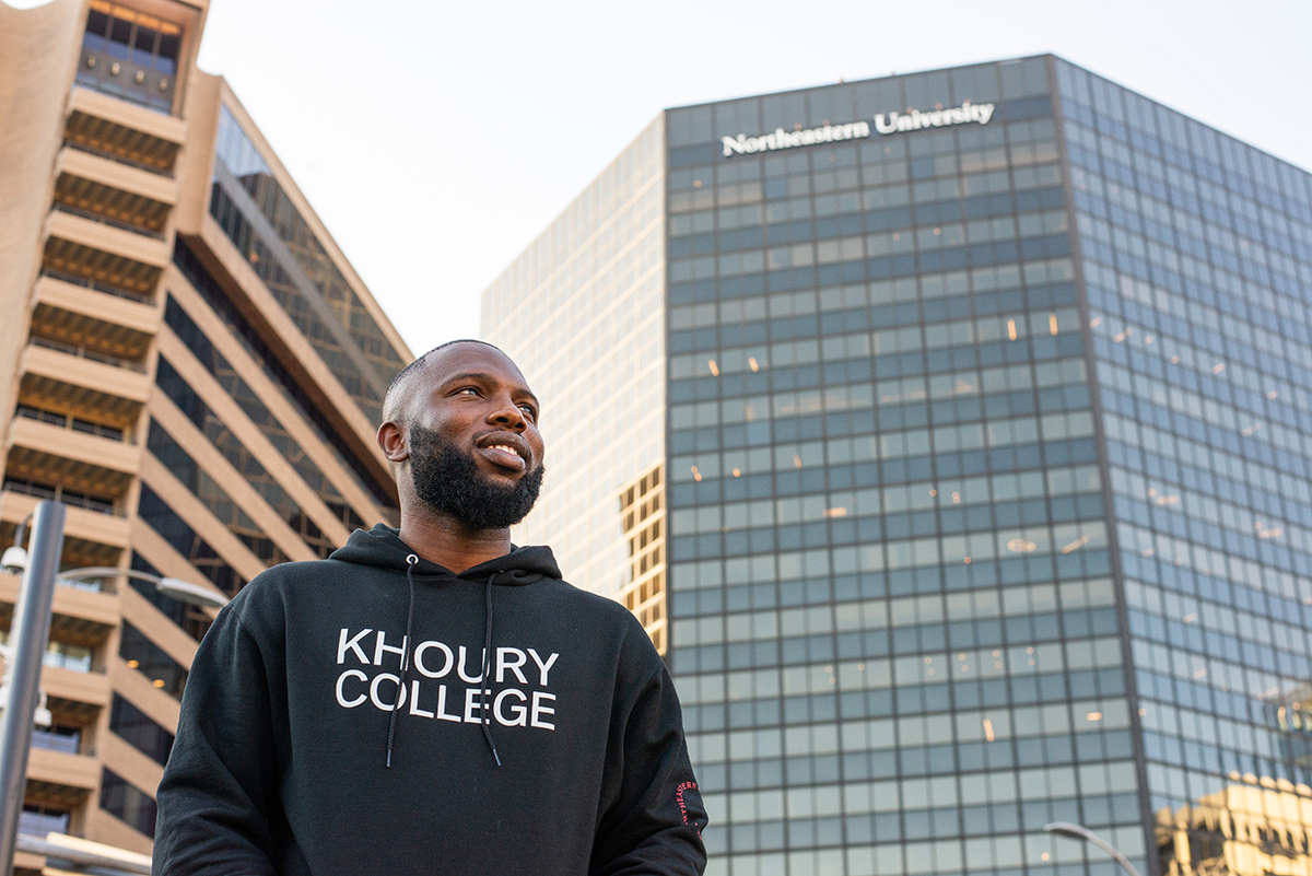 A student wearing a Khoury College sweatshirt stands in front of the Northeastern Arlington building