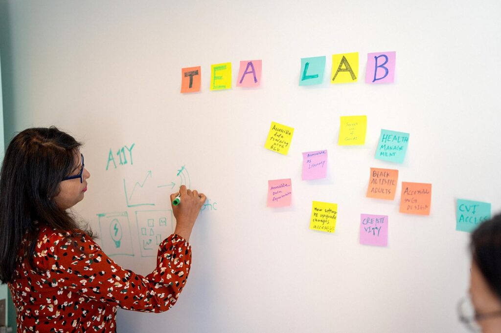 Maitraye Das writes on a whiteboard with a marker. There are several yellow, orange, and pink Post-it notes attached to the whiteboard.