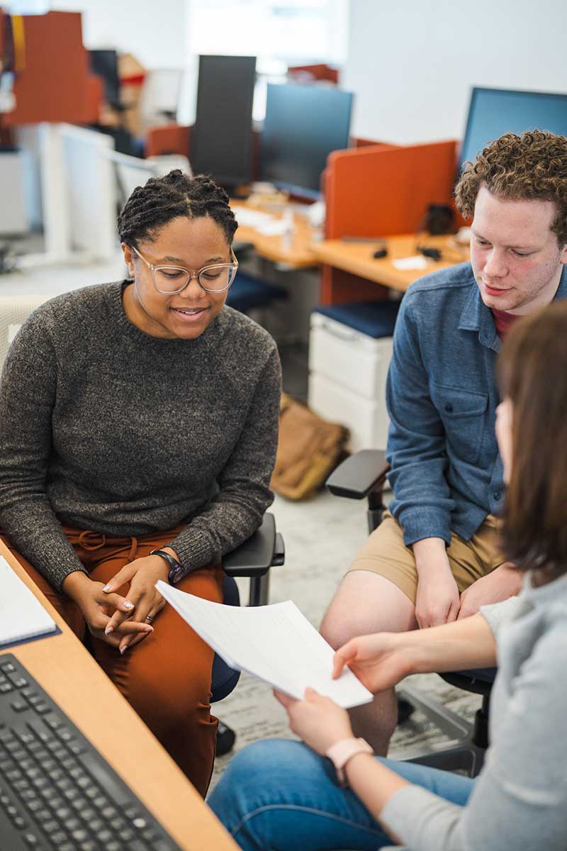 Three students sitting in a computer lab discuss a project. One student is holding a stack of papers in her hand.