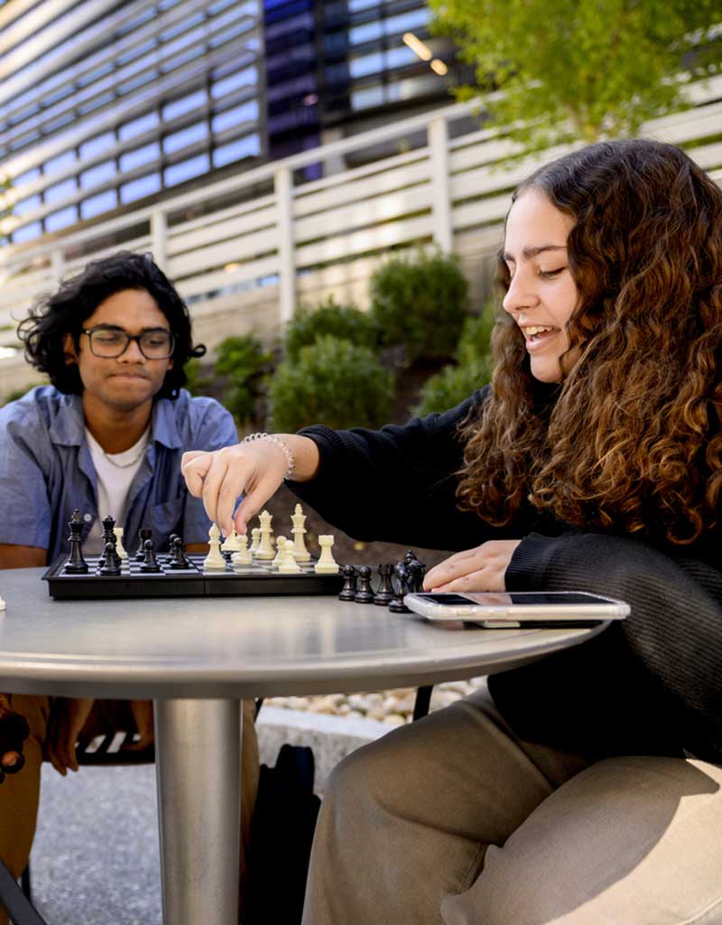 Two students sit outdoors playing chess on a round table. One student is moving a white piece.