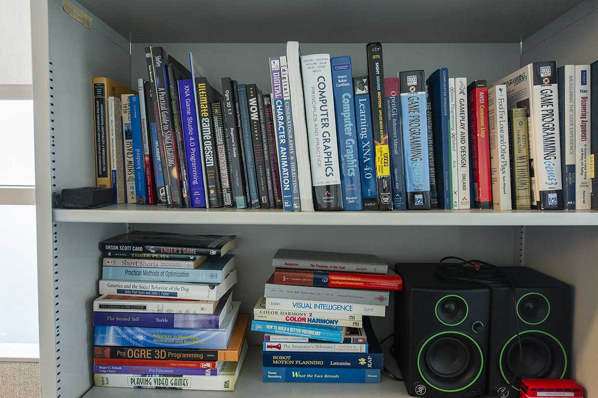 A view of two shelves in a Khoury research lab. The shelves hold dozens of game design and computer graphics books and two audio speakers.